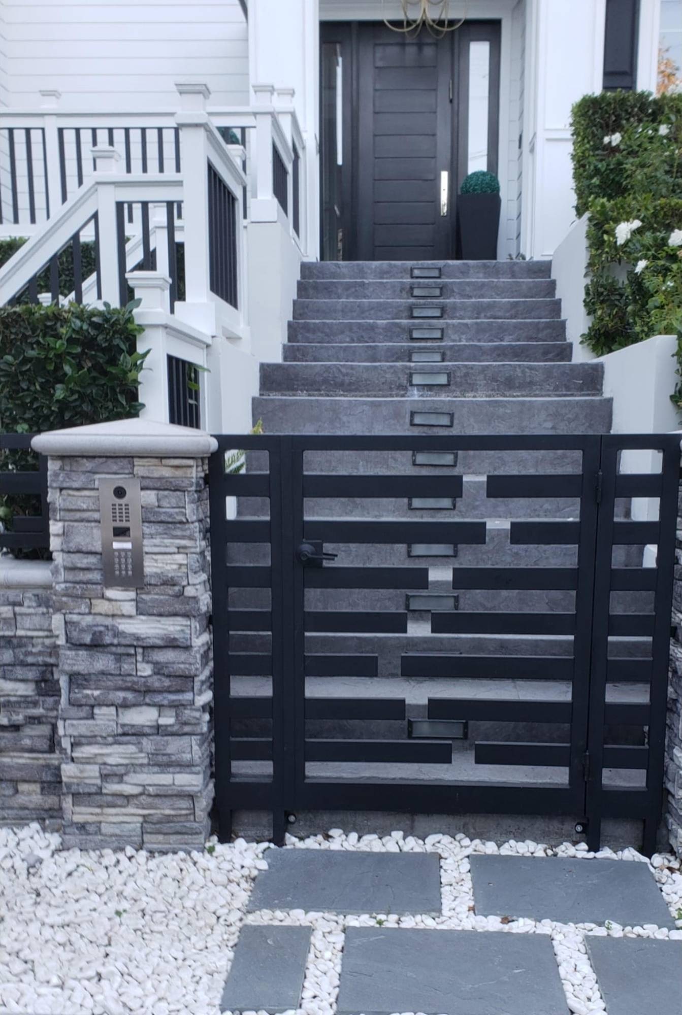 Modern black pedestrian gate with offset horizontal bars beside a stone pillar with intercom and keypad