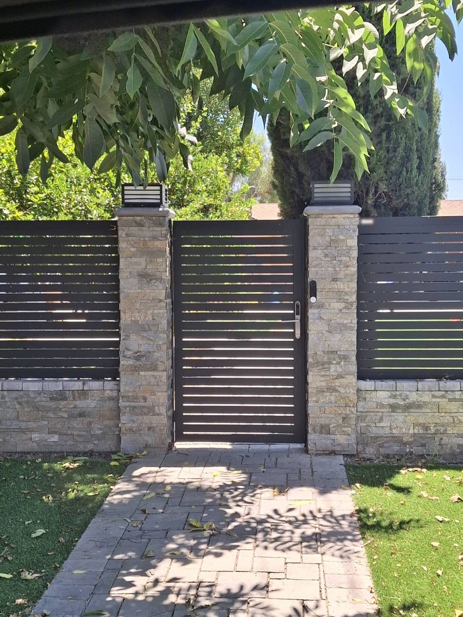 Black horizontal slat pedestrian gate between stacked stone pillars with electronic lock and matching slat fence