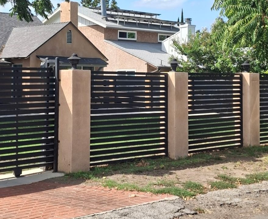 Tan stucco-finished pillars with lamp caps supporting a black slat fence and driveway gate